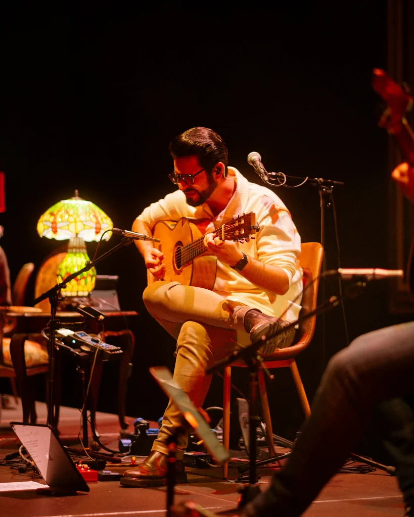 Guitarrista do Luís Trigacheiro a tocar no palco, com luz quente, no concerto no Quartel das Artes em 2025 — fotografia por xpectrum. [NOTA: confirma se o instrumento na foto é mesmo harmónica — posso ajustar para "flauta", "ocarina" ou outro se quiseres.]