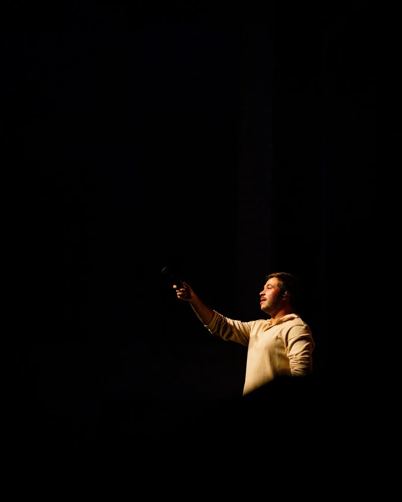 Luís Trigacheiro com o braço estendido a apontar o microfone para o público, iluminado por um foco isolado no palco escuro do Quartel das Artes em 2025 — xpectrum fotografia de concertos.