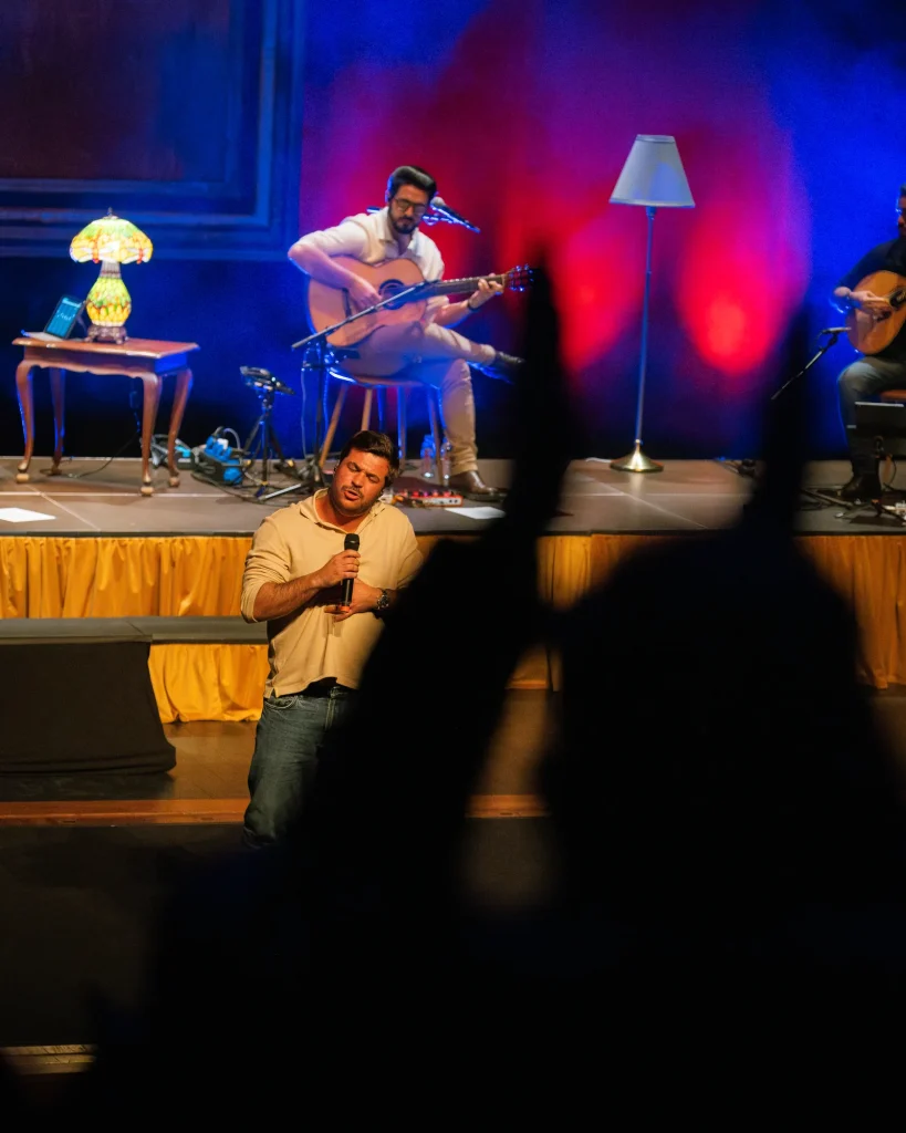 Luís Trigacheiro a cantar. Vista do público com a banda a acompanhar no palco ao fundo, silhuetas da plateia em primeiro plano, no Quartel das Artes em 2025 — fotografia por xpectrum.