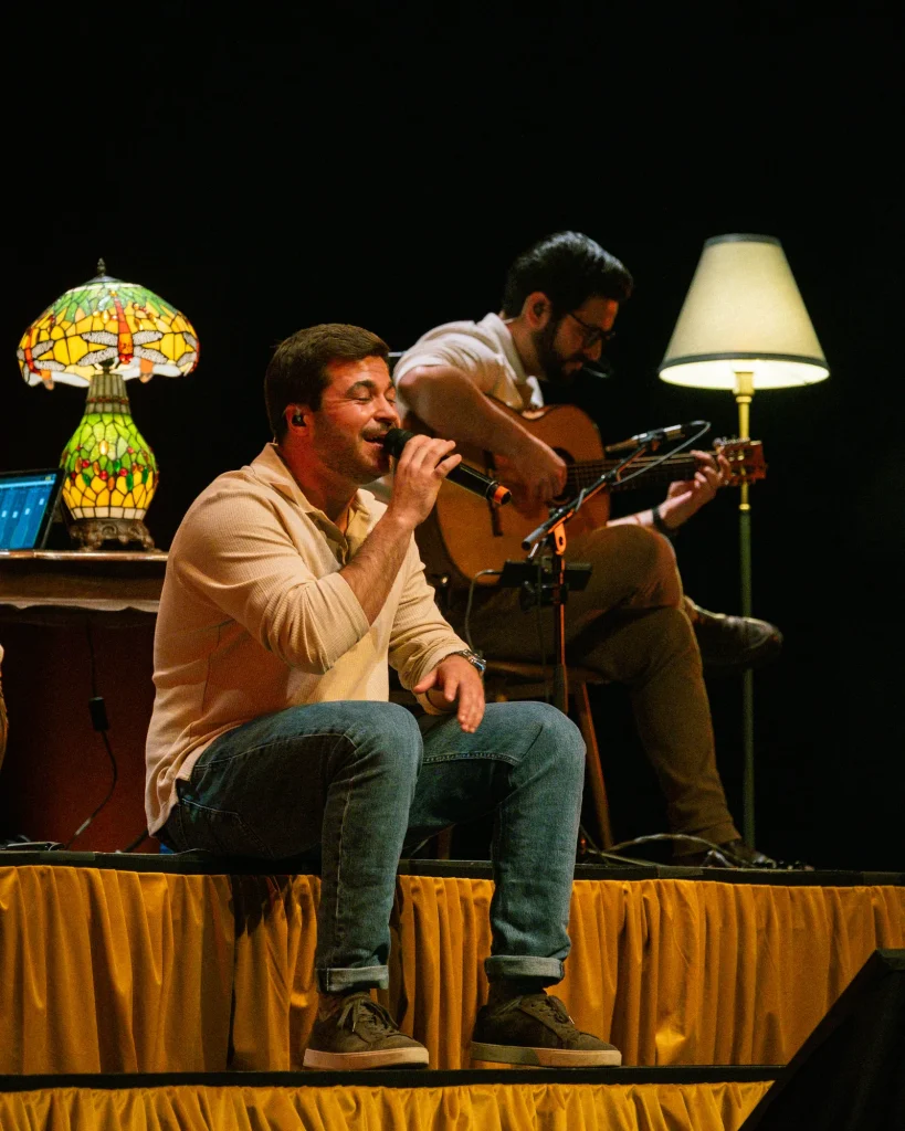 Luís Trigacheiro sentado a cantar ao microfone com guitarrista português a acompanhar ao fundo, candeeiro à esquerda no palco do Quartel das Artes em 2025 — fotografia por xpectrum.