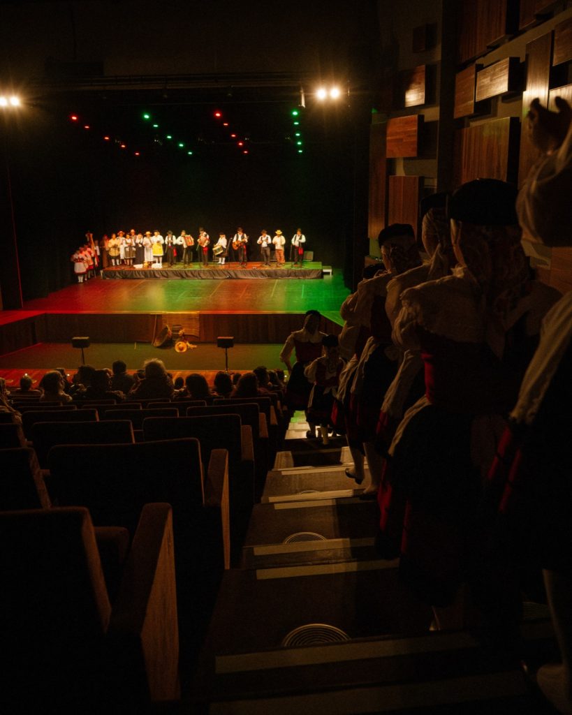 Vista geral da sala do Quartel das Artes com plateia, palco iluminado e rancho folclórico em atuação durante o evento MOB 2025