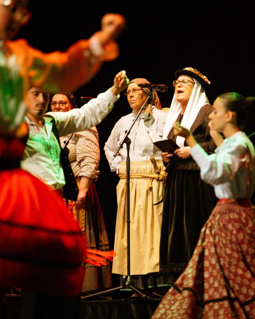 Cantadeiras de rancho folclórico a cantar ao microfone rodeadas de dançarinos em trajes tradicionais no evento MOB 2025 no Quartel das Artes