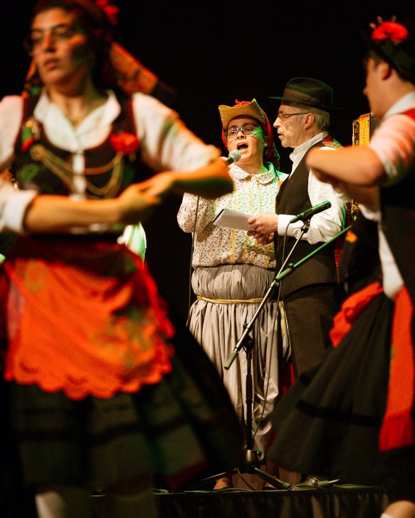 Vocalista de rancho folclórico em traje tradicional português a cantar ao microfone rodeado de dançarinos no evento MOB 2025 no Quartel das Artes