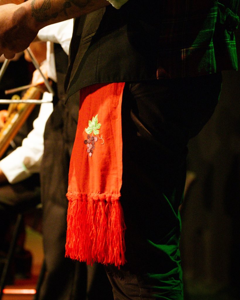 Detalhe de lenço vermelho com bordado de cacho de uvas no colete preto de traje folclórico masculino no evento MOB 2025 no Quartel das Artes