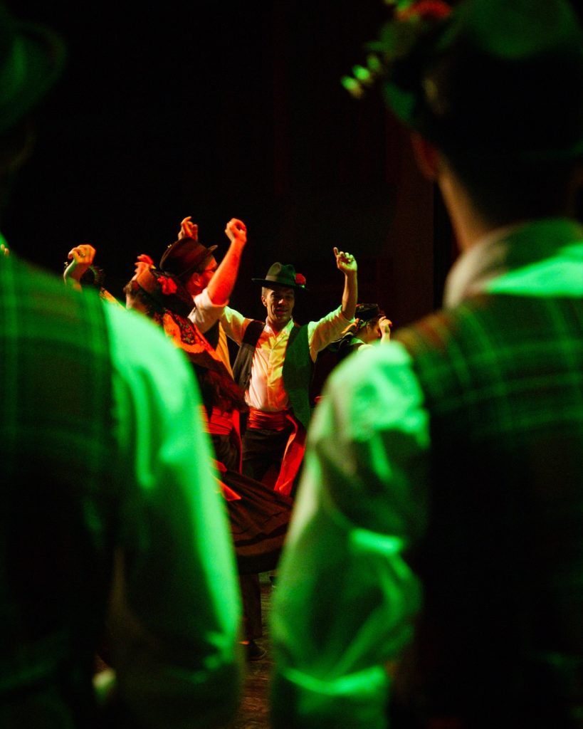 Dançarino de rancho folclórico em destaque no centro do palco iluminado a verde, enquadrado entre outros bailarinos, no evento MOB 2025 no Quartel das Artes