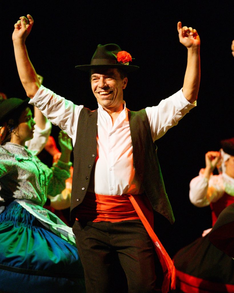 Bailarino de rancho folclórico com chapéu tradicional e flor vermelha a sorrir com os braços levantados no palco do evento MOB 2025 no Quartel das Artes