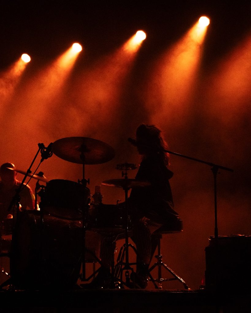 Sangue Suor ao vivo no Quartel das Artes 2025 – silhueta da baterista com focos laranja – fotografia de concertos xpectrum