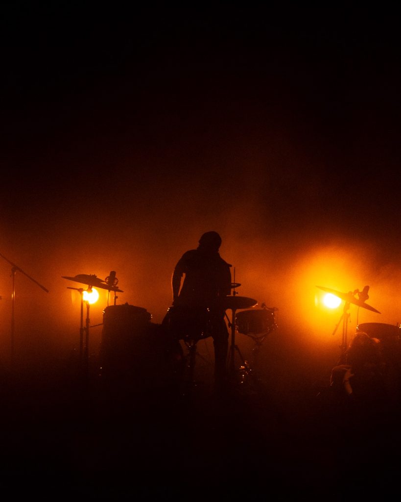 Sangue Suor ao vivo no Quartel das Artes 2025 – baterista em silhueta com luz laranja e fumo – fotografia de concertos xpectrum