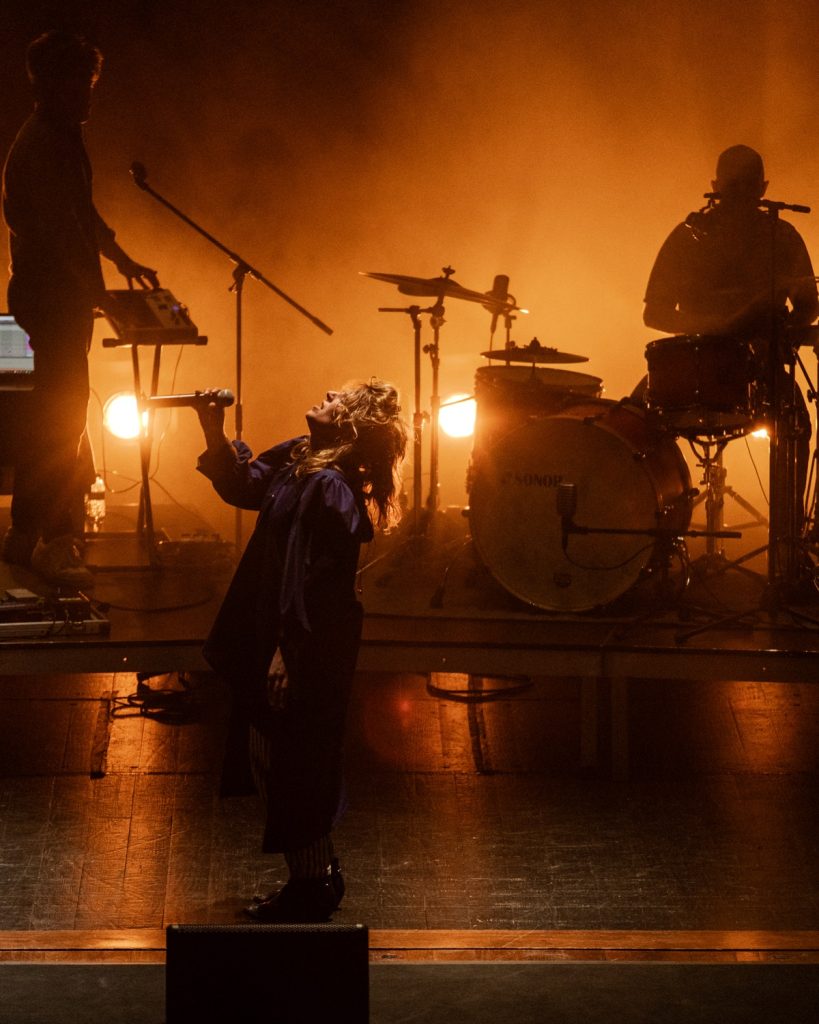 Sangue Suor no Quartel das Artes 2025 – vocalista curvada no palco com microfone e luz laranja – fotografia de concertos xpectrum