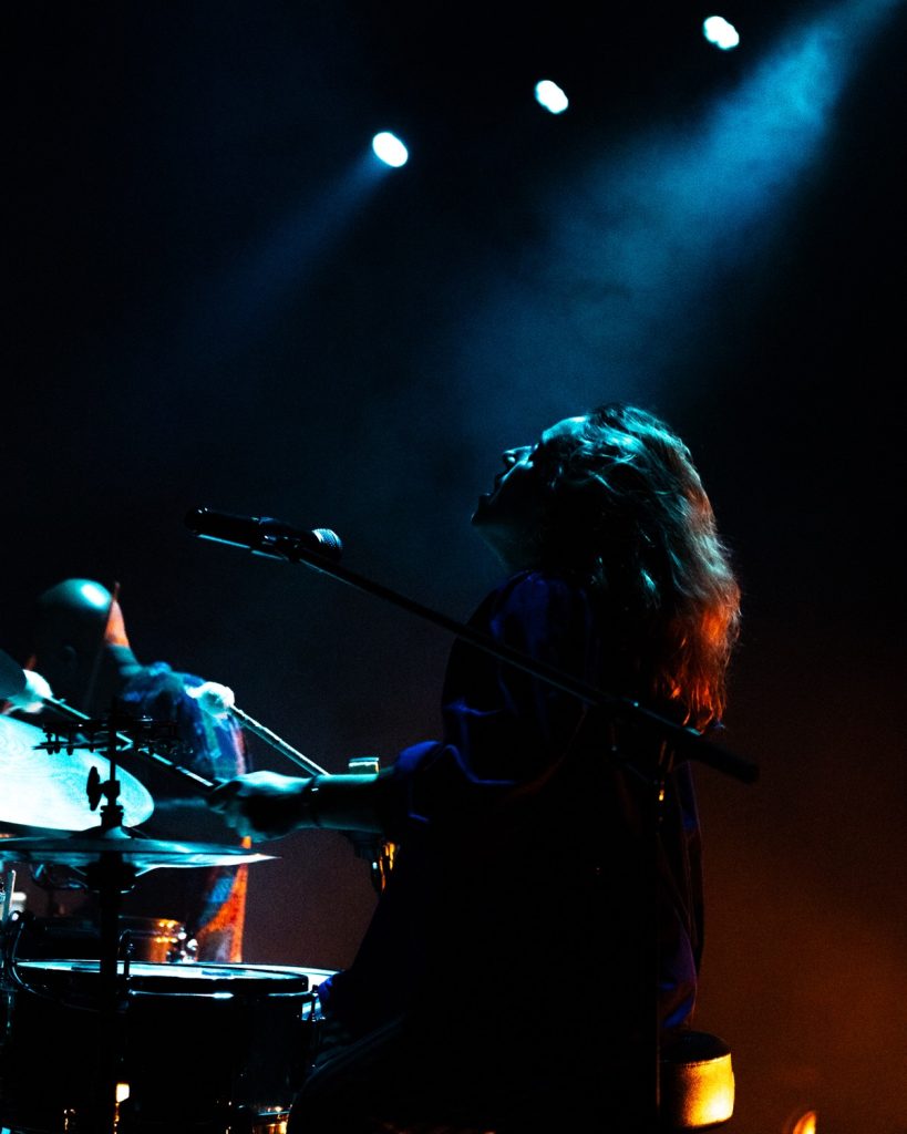 Sangue Suor ao vivo no Quartel das Artes 2025 – baterista iluminada por luz azul e focos brancos – fotografia de concertos xpectrum