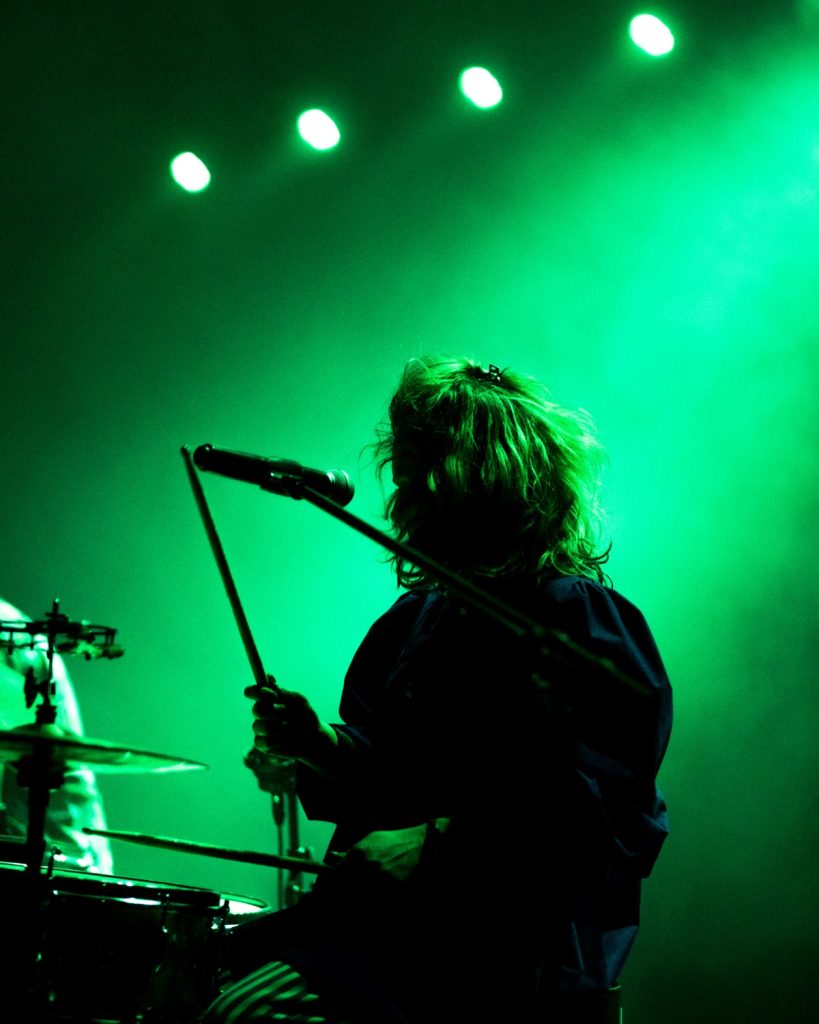 Sangue Suor ao vivo no Quartel das Artes 2025 – silhueta da baterista com luz verde – fotografia de concertos xpectrum