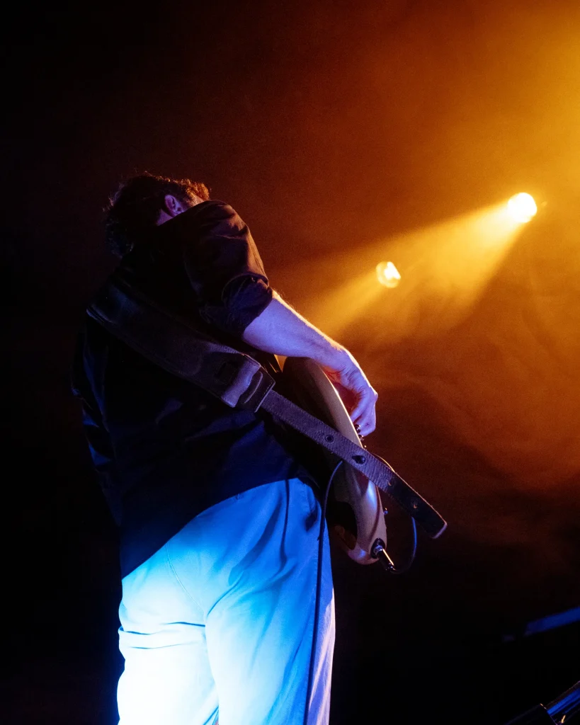 Baixista da banda Solaris de lado com baixo elétrico, iluminado por luz dourada dramática em palco com fumo, Quartel das Artes 2025 — xpectrum