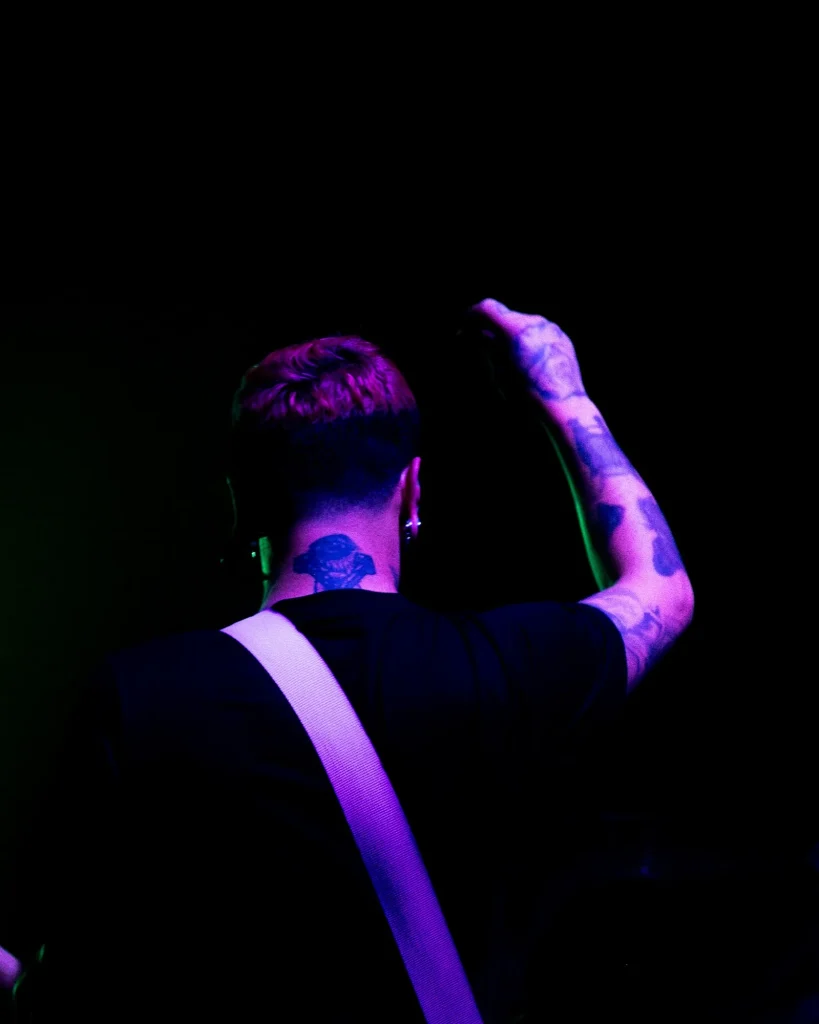 Pormenor do baixista dos Avesso de costas com tatuagens nos braços e cabelo cor-de-rosa sob luz roxa no Quartel das Artes em 2025, fotografia de concertos xpectrum.