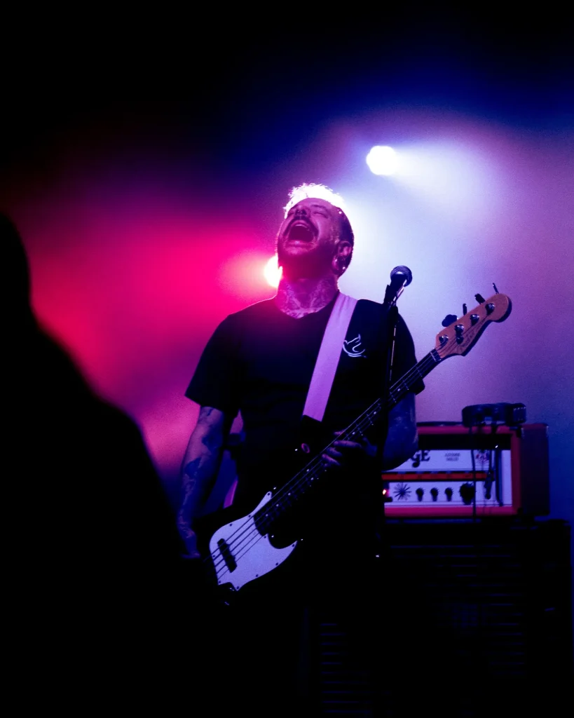 Baixista dos Avesso a gritar ao microfone com cabeça erguida sob luz rosa e azul no Quartel das Artes em 2025, fotografia de concertos xpectrum.