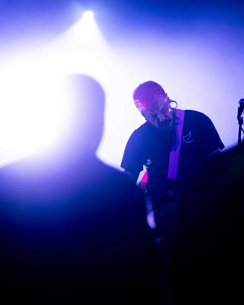 Baixista e vocalista dos Avesso de cabeça baixa a tocar baixo com fundo iluminado por contraluz violeta no Quartel das Artes em 2025, fotografia de concertos xpectrum.