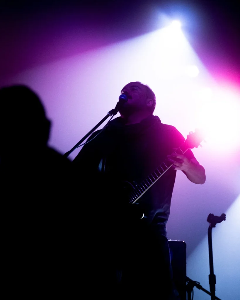 Guitarrista dos Avesso visto em contrapicado a cantar ao microfone com luzes rosa e azul no Quartel das Artes em 2025, fotografia de concertos xpectrum.