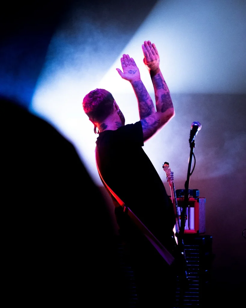Baixista e vocalista dos Avesso de braços erguidos com tatuagens visíveis sob luz azul e fumo no Quartel das Artes em 2025, fotografia de concertos xpectrum.