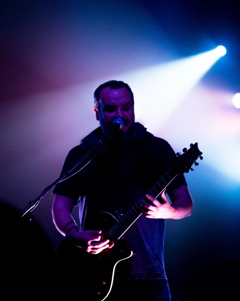 Guitarrista dos Avesso a cantar ao microfone enquanto toca guitarra elétrica com feixes de luz azul ao fundo no Quartel das Artes em 2025, fotografia de concertos xpectrum.