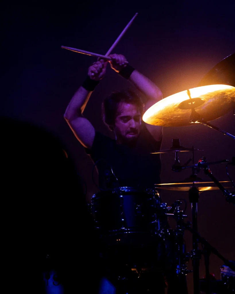 Baterista dos Avesso com baquetas no ar prestes a bater num prato iluminado a laranja no concerto do Quartel das Artes em 2025, fotografia de concertos xpectrum.