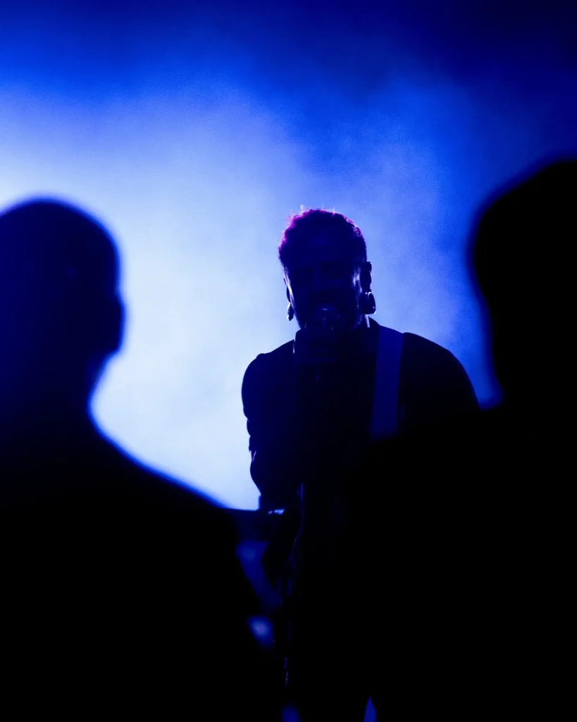 Vocalista dos Avesso ao microfone visto entre silhuetas do público sob luz azul no Quartel das Artes em 2025, fotografia de concertos xpectrum.
