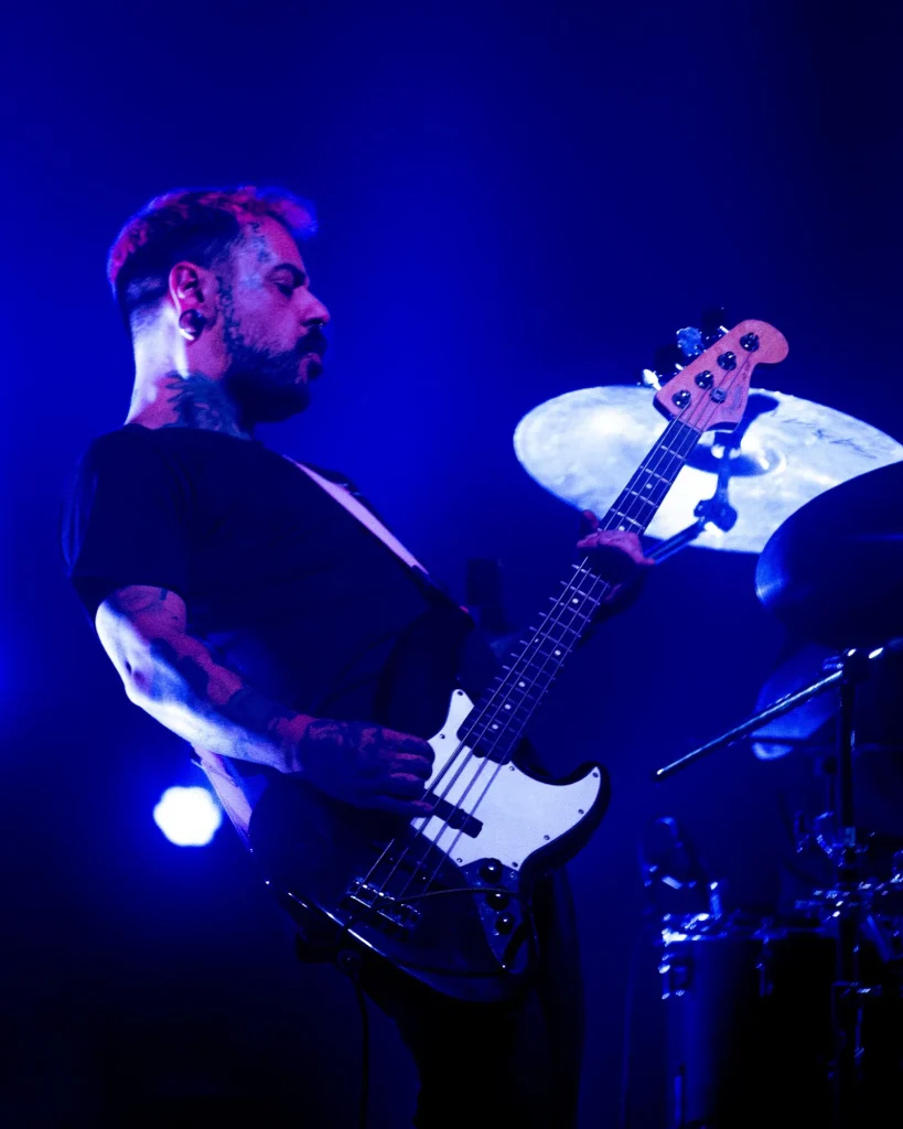 Baixista dos Avesso com tatuagens nos braços a tocar baixo branco e preto sob luz azul no Quartel das Artes em 2025, fotografia de concertos xpectrum.