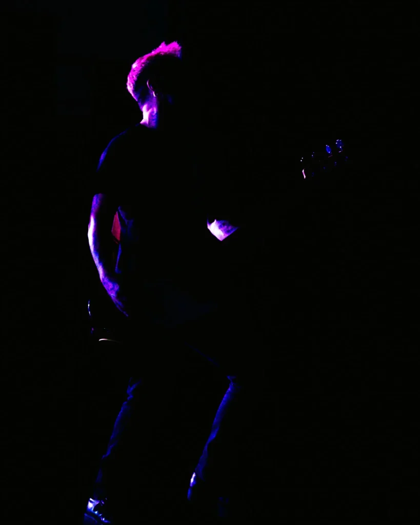 Baixista dos Avesso recortado em silhueta com cabelo iluminado a magenta no escuro do palco do Quartel das Artes em 2025, fotografia de concertos xpectrum.