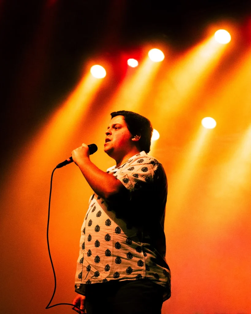 Vocalista dos Bandidos do Cante de camisa estampada a cantar ao microfone sob feixes de luz amarelos e laranjas no Quartel das Artes em 2025, fotografia de concertos xpectrum.