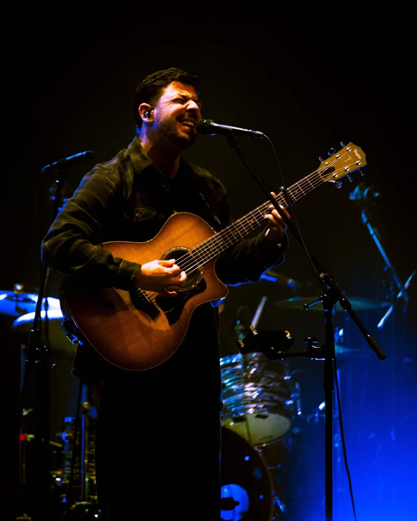 Vocalista e guitarrista dos Bandidos do Cante a cantar ao microfone enquanto toca guitarra acústica, com bateria iluminada a azul ao fundo, ao vivo no Quartel das Artes em 2025, fotografia de concertos xpectrum.