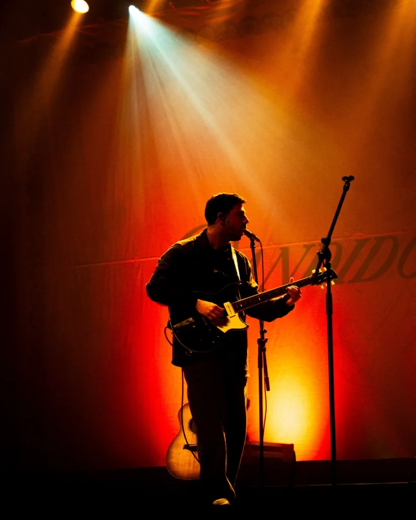 Silhueta de músico dos Bandidos do Cante com guitarra elétrica em contraluz laranja sobre o cenário no Quartel das Artes em 2025, fotografia de concertos xpectrum.
