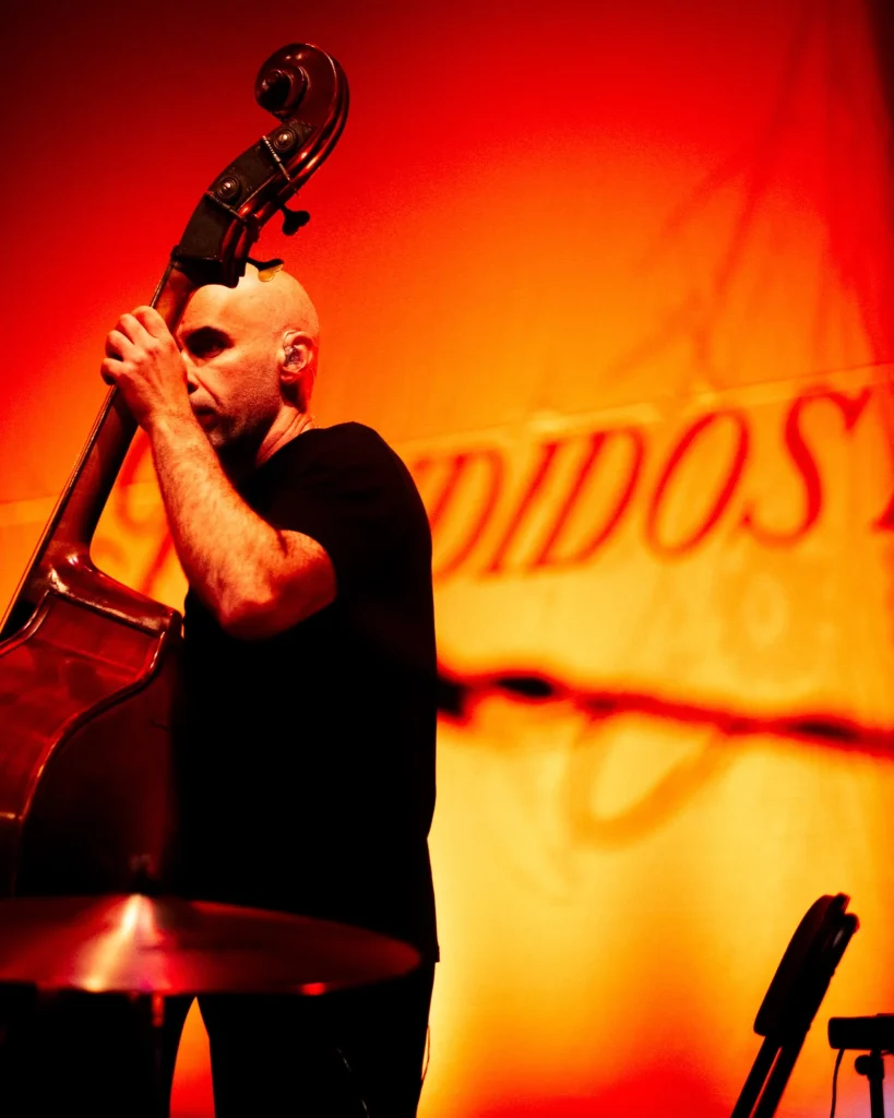 Contrabaixista dos Bandidos do Cante a tocar contrabaixo acústico sobre fundo vermelho e dourado com o nome da banda no Quartel das Artes em 2025, fotografia de concertos xpectrum.