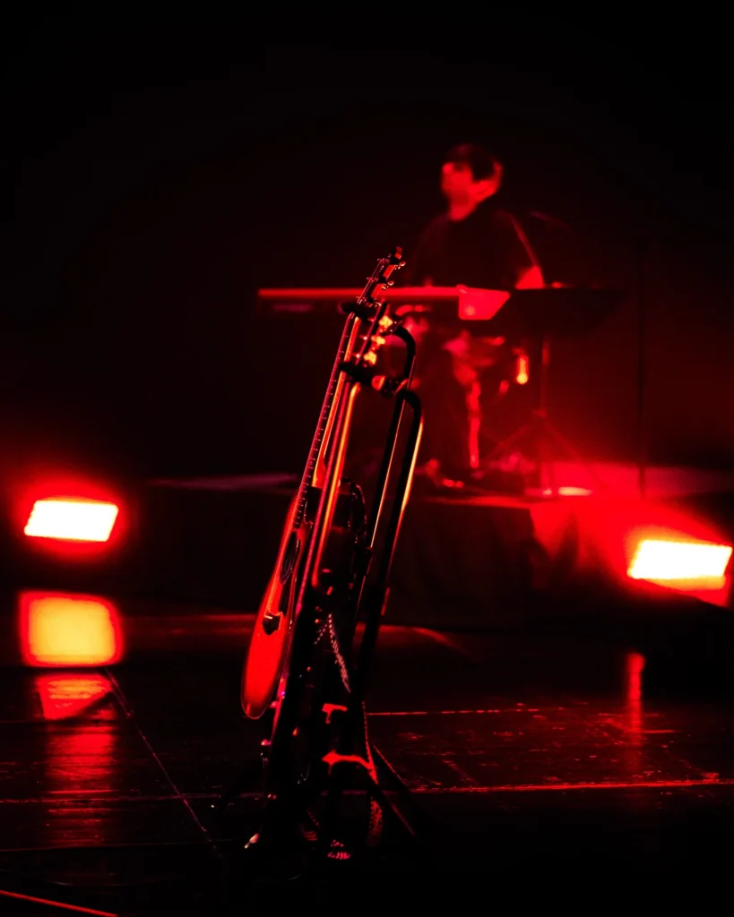 Pormenor de guitarras em suporte no palco com tecladista desfocado ao fundo sob luz vermelha intensa, concerto Bandidos do Cante no Quartel das Artes em 2025, fotografia de concertos xpectrum.