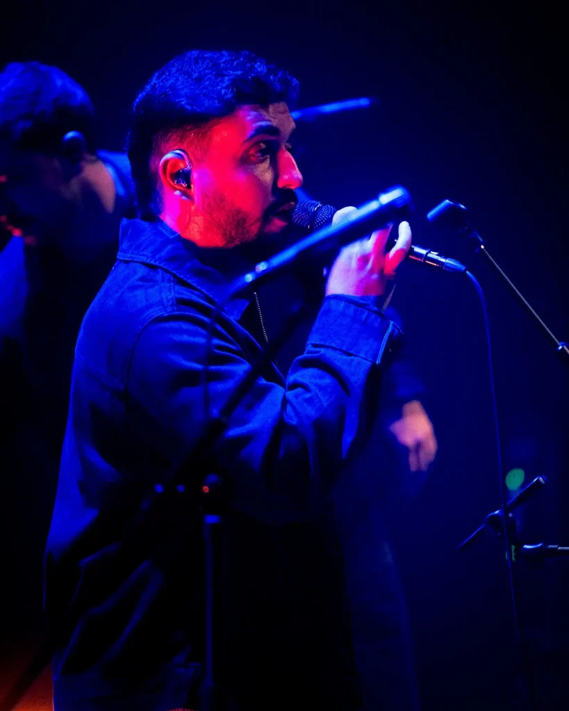 Vocalista dos Bandidos do Cante de jaqueta de ganga a cantar ao microfone sob luz azul e vermelha no Quartel das Artes em 2025, fotografia de concertos xpectrum.