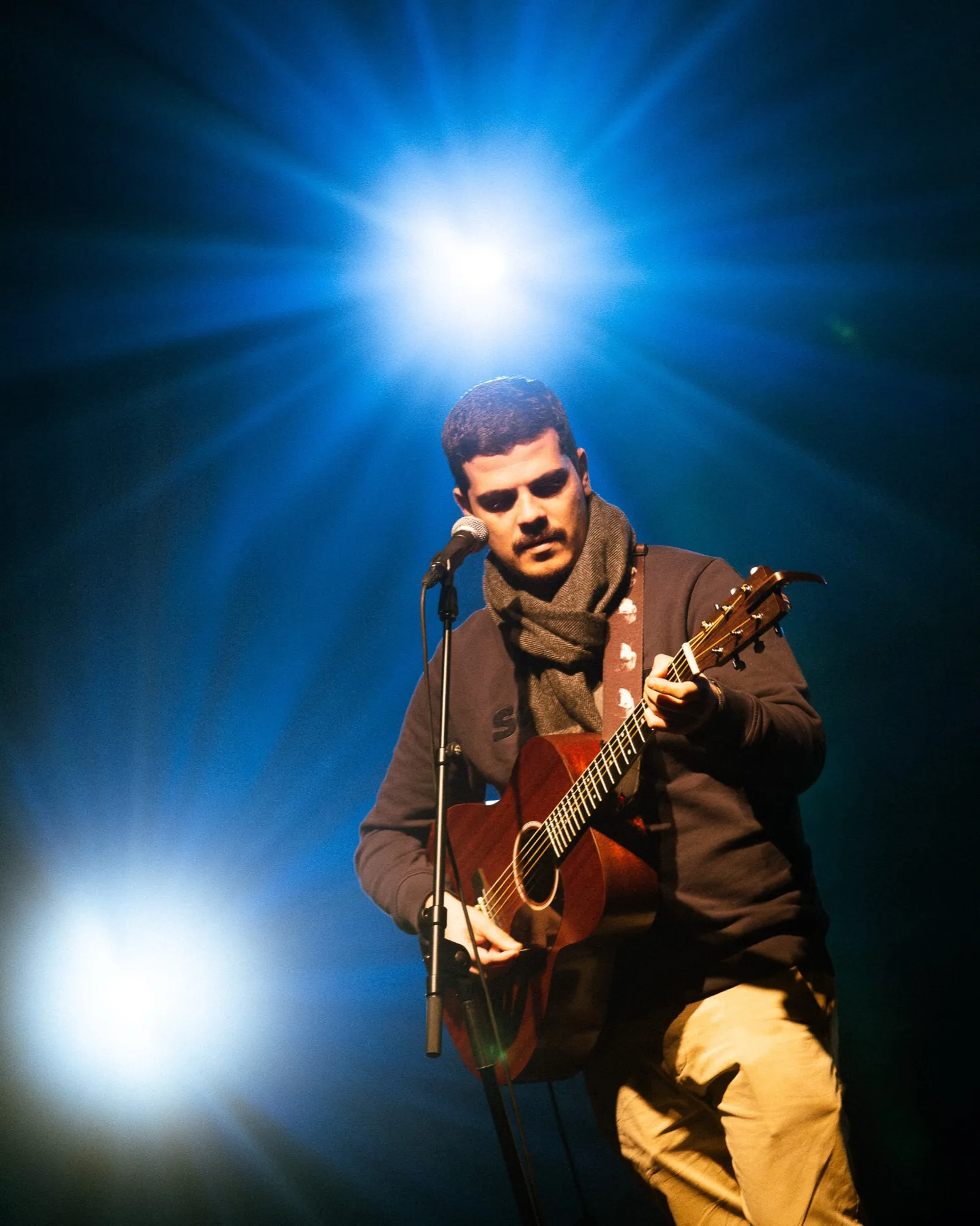 Vocalista dos Duque Província de cachecol bege a tocar guitarra acústica ao microfone sob estrela de luz azul no Quartel das Artes em 2025, fotografia de concertos xpectrum.