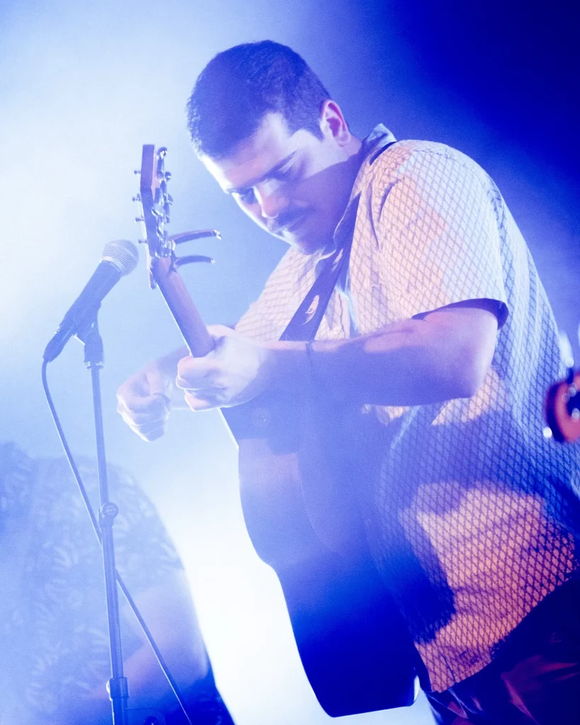 Guitarrista e vocalista dos Duque Província de camisa branca padronizada a tocar guitarra acústica em close sob luz azul no Quartel das Artes em 2025, fotografia de concertos xpectrum.