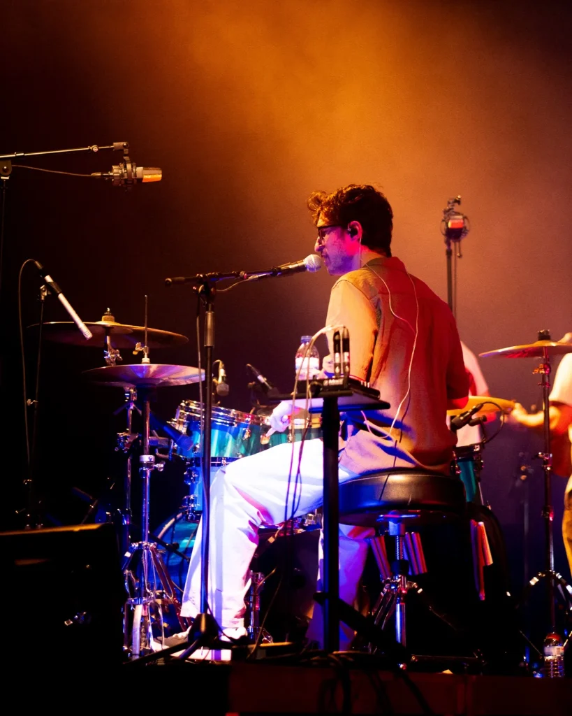 Baterista dos Duque Província sentado a cantar ao microfone com bateria ao lado e luz quente laranja no Quartel das Artes em 2025, fotografia de concertos xpectrum.