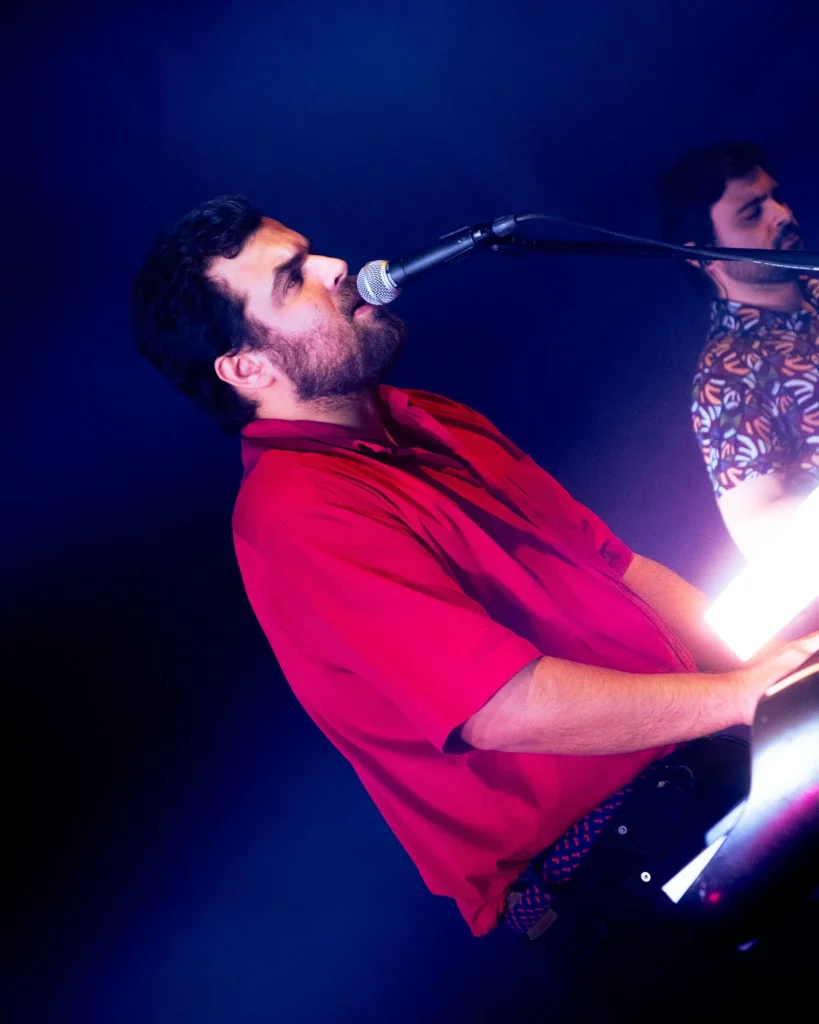 Vocalista e teclista dos Duque Província de camisa vermelha em contrapicado a cantar ao microfone com colega ao fundo sob luz roxa no Quartel das Artes em 2025, fotografia de concertos xpectrum.
