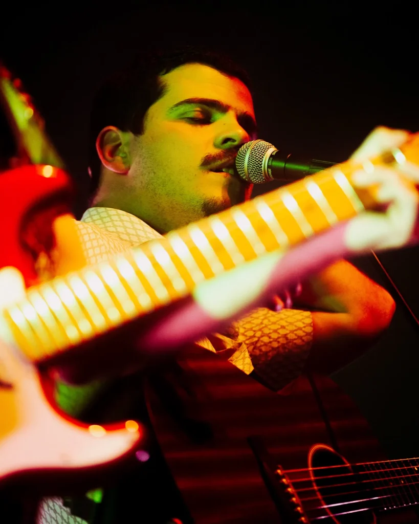 Vocalista dos Duque Província a cantar ao microfone com guitarras desfocadas em primeiro plano sob luz verde e laranja no Quartel das Artes em 2025, fotografia de concertos xpectrum.