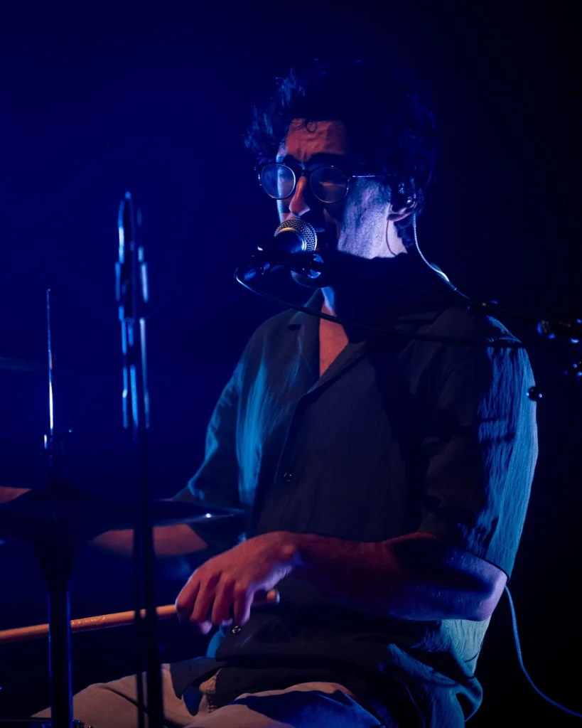 Baterista dos Duque Província de óculos a cantar ao microfone com pratos de bateria à frente e luz azul no Quartel das Artes em 2025, fotografia de concertos xpectrum.