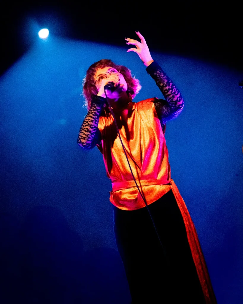 Vocalista dos Equinócio a cantar ao microfone com colete cetim laranja sob luz azul de palco no Quartel das Artes 2025