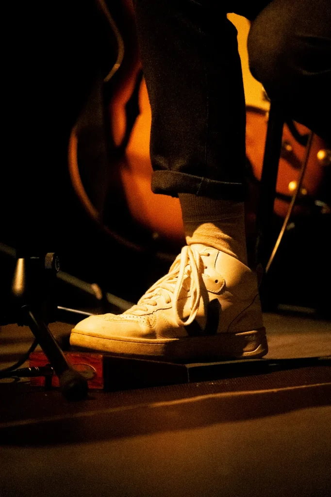 Pormenor de ténis branco do músico em palco com guitarra acústica desfocada no concerto dos Equinócio no Quartel das Artes 2025