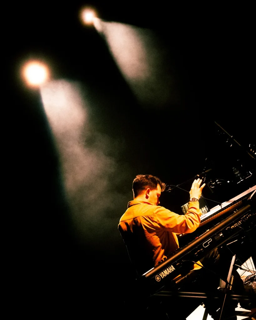 Tecladista dos Equinócio de costas ao teclado Yamaha sob feixes de luz e fumo de palco no Quartel das Artes 2025
