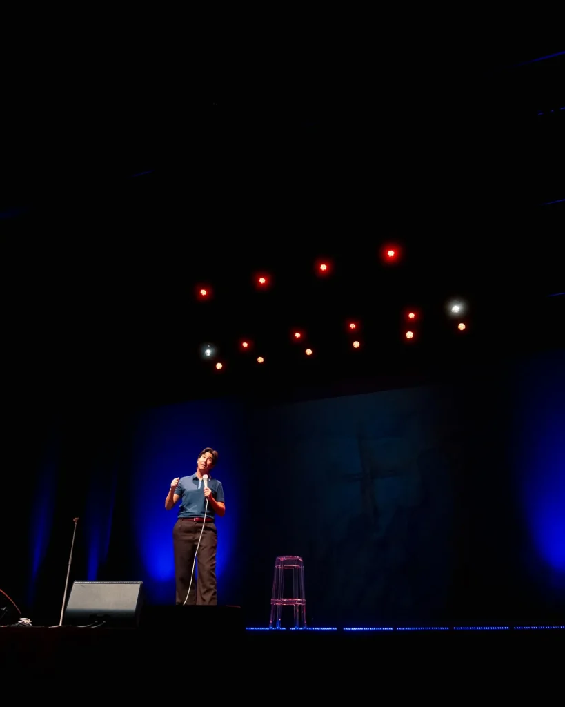 Plano largo de Luana do Bem em palco sob a vara de luzes vermelhas e brancas do Quartel das Artes em 2025, banco transparente e monitor a seu lado — fotografia de espetáculo de humor por xpectrum.