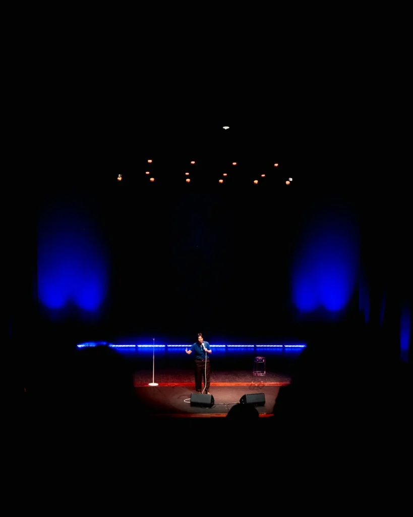Vista geral do palco do Quartel das Artes em 2025 com Luana do Bem ao centro, recortada pela linha azul do palco e pelas luzes da teia — fotografia de espetáculo de humor por xpectrum.