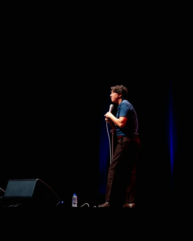 Luana do Bem de perfil iluminada por um foco quente em palco escurecido do Quartel das Artes em 2025, microfone na mão e cortina azul ao fundo — fotografia de espetáculo de humor por xpectrum.