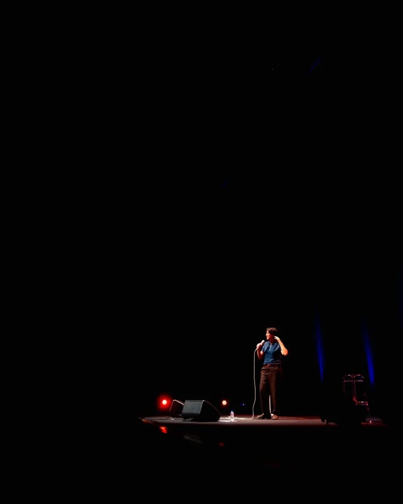 Plano aberto de Luana do Bem isolada no canto direito do palco do Quartel das Artes em 2025, monitor de retorno e refletores vermelhos a desenhar a cena — fotografia de espetáculo de humor por xpectrum.