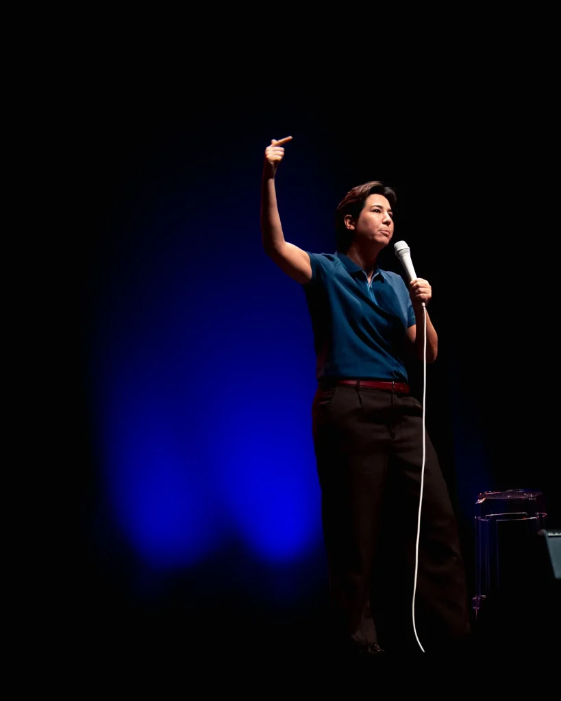 Luana do Bem em palco de pólo azul aponta o dedo para cima com o microfone na mão direita, iluminação azul a recortar a silhueta no Quartel das Artes em 2025 — fotografia de espetáculo de humor por xpectrum.