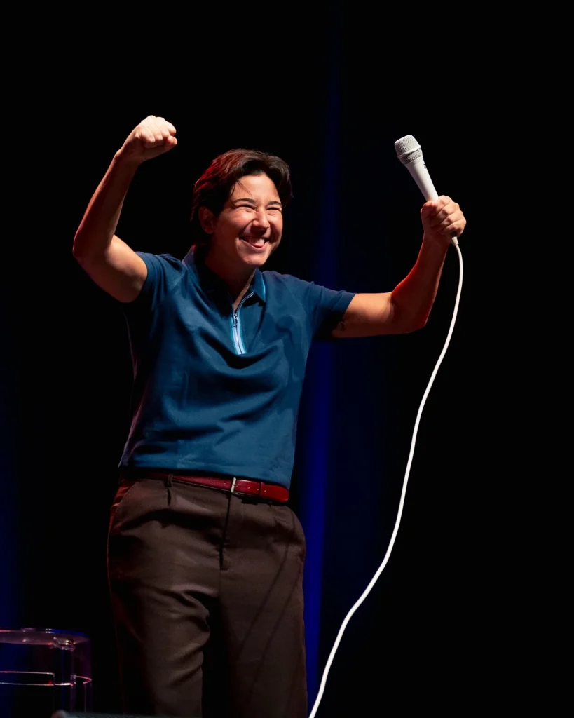 Luana do Bem celebra com um punho erguido e o microfone esticado no ar, gargalhada em palco no Quartel das Artes em 2025 — fotografia de espetáculo de humor por xpectrum.