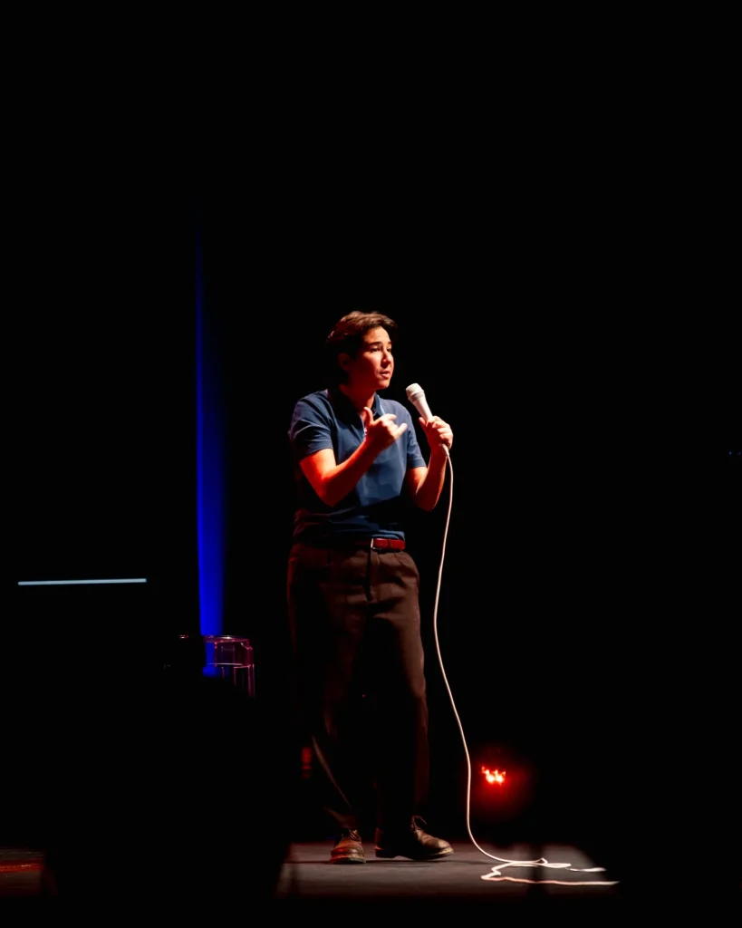Luana do Bem a contar uma piada gesticulando com a mão livre, microfone aceso e luz vermelha no chão do palco do Quartel das Artes em 2025 — fotografia de espetáculo de humor por xpectrum.