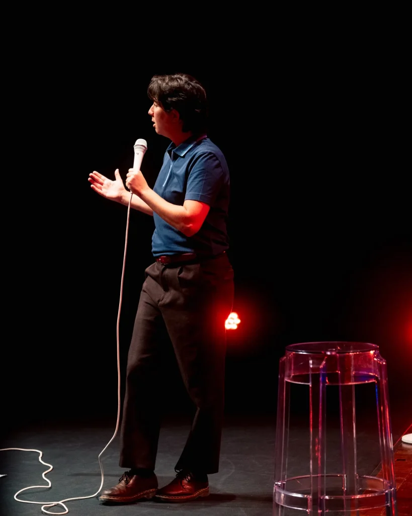 Luana do Bem de perfil em palco a falar ao microfone, banco transparente e refletor encarnado em segundo plano no Quartel das Artes em 2025 — fotografia de espetáculo de humor por xpectrum.