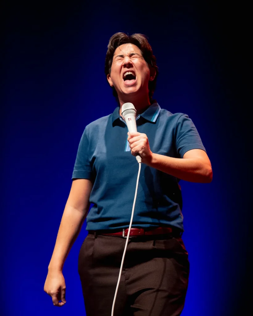 Luana do Bem grita ao microfone com expressão exagerada e olhos cerrados, fundo azul intenso no palco do Quartel das Artes em 2025 — fotografia de espetáculo de humor por xpectrum.